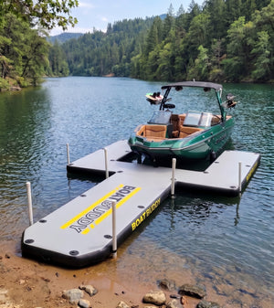 An inflatable floating boat dock and jetty system on a lake, with a green and white wake boat parked on the platform. The pontoon has a gray and white design with yellow and black branding text.