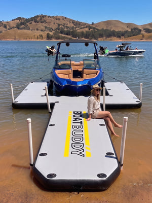 A woman sits on the front platform of a white and yellow inflatable boat buddy floating U dock, jetty and pontoon, with a wake boat moored behind it on a lake with hills in the background.